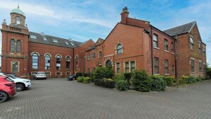 A large brick building with a clock tower, a paved courtyard, and parked cars.