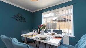 A dining area with a white table set for four, blue chairs, and a window with blinds. - Spacious 3 Bedroom Home in Guisborough (Redcar and Cleveland)