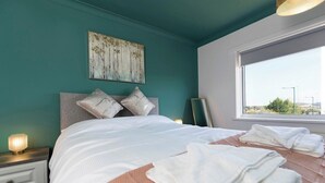 A neatly made bed with white linens and a pink accent, a bedside table with a lamp, and a window with a view of the outside. - Spacious 3 Bedroom Home in Guisborough (Redcar and Cleveland)