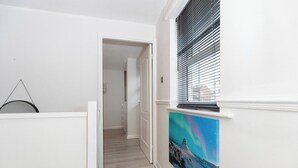 Um corredor com piso de madeira, paredes brancas, uma porta que dá para outro quarto e uma janela com persianas.