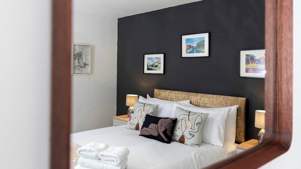 Cama bem arrumada com lençóis brancos e almofadas decorativas, mesa de cabeceira com abajur e quadros nas paredes.