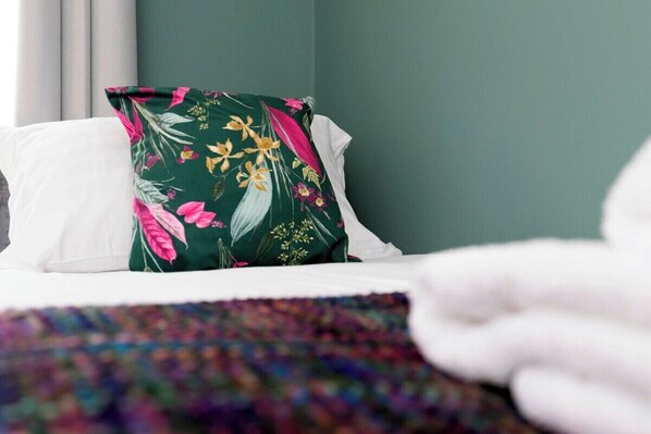 A bed with white linens, a green pillow with floral patterns, and a multicolored blanket.