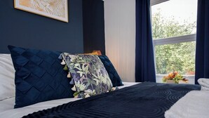 Ein ordentlich bezogenes Bett mit einem dunkelblauen Quilt und Kissen, ein gerahmtes Kunstwerk an der Wand und ein Fenster mit Blick auf Grünflächen.