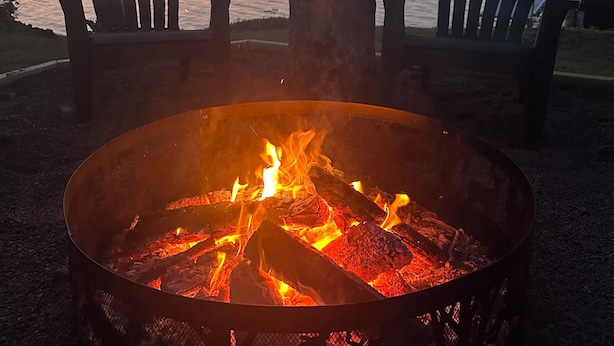 Ein Lagerfeuer mit zwei Adirondack-Stühlen, die auf einen See bei Sonnenuntergang blicken.