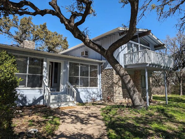 Une maison à deux étages, avec des fondations en pierre, un balcon et un grand arbre à l’avant.