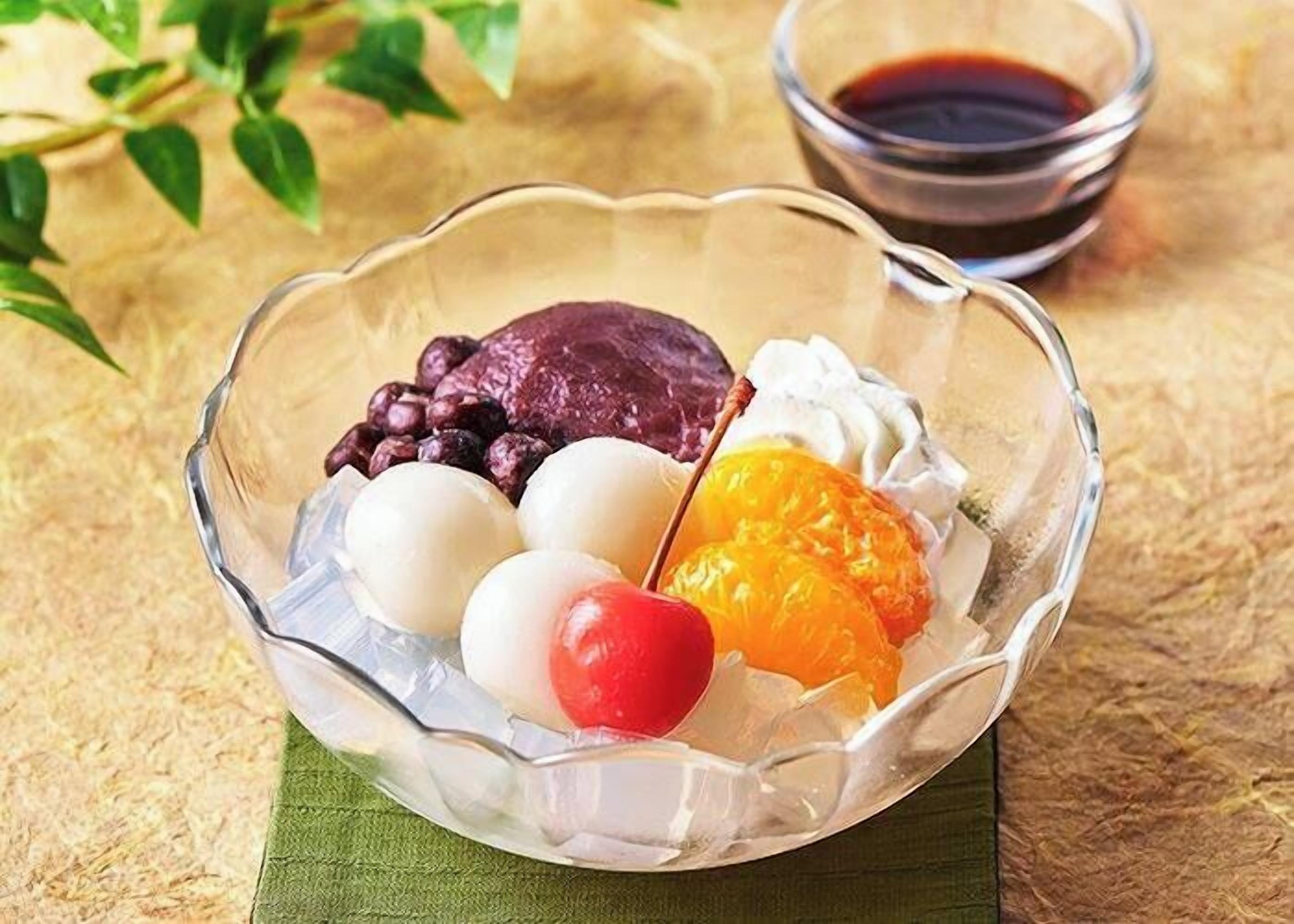 A clear glass bowl containing various food items, a small bowl of dark sauce, and a green napkin.