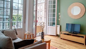 Een woonkamer met een bank, een salontafel, een televisie op een houten standaard, een ronde spiegel aan de muur en een deur die leidt naar een buitenbalkon.