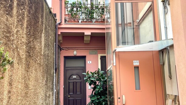 Une ruelle étroite avec un immeuble dont la porte est d’un brun foncé, un balcon orné de plantes en pot et une structure en bois sur la droite.