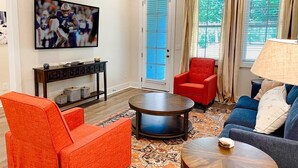 A living room with a red armchair, blue sofa, and a round coffee table. There is a television mounted on the wall and a window with curtains. - Aubie’s Favorite Place near Campus and Tiger Town with 2 King & 2 Queen beds  (Auburn)