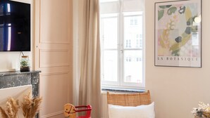Een kamer met een houten schommelstoel, een witte stoel, een vaas met gedroogde planten, botanische kunst in een lijst en een televisie aan de muur.