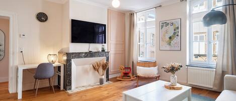 A living room with a fireplace, a TV mounted on the wall, a dining table, and a view of the outside through a window.