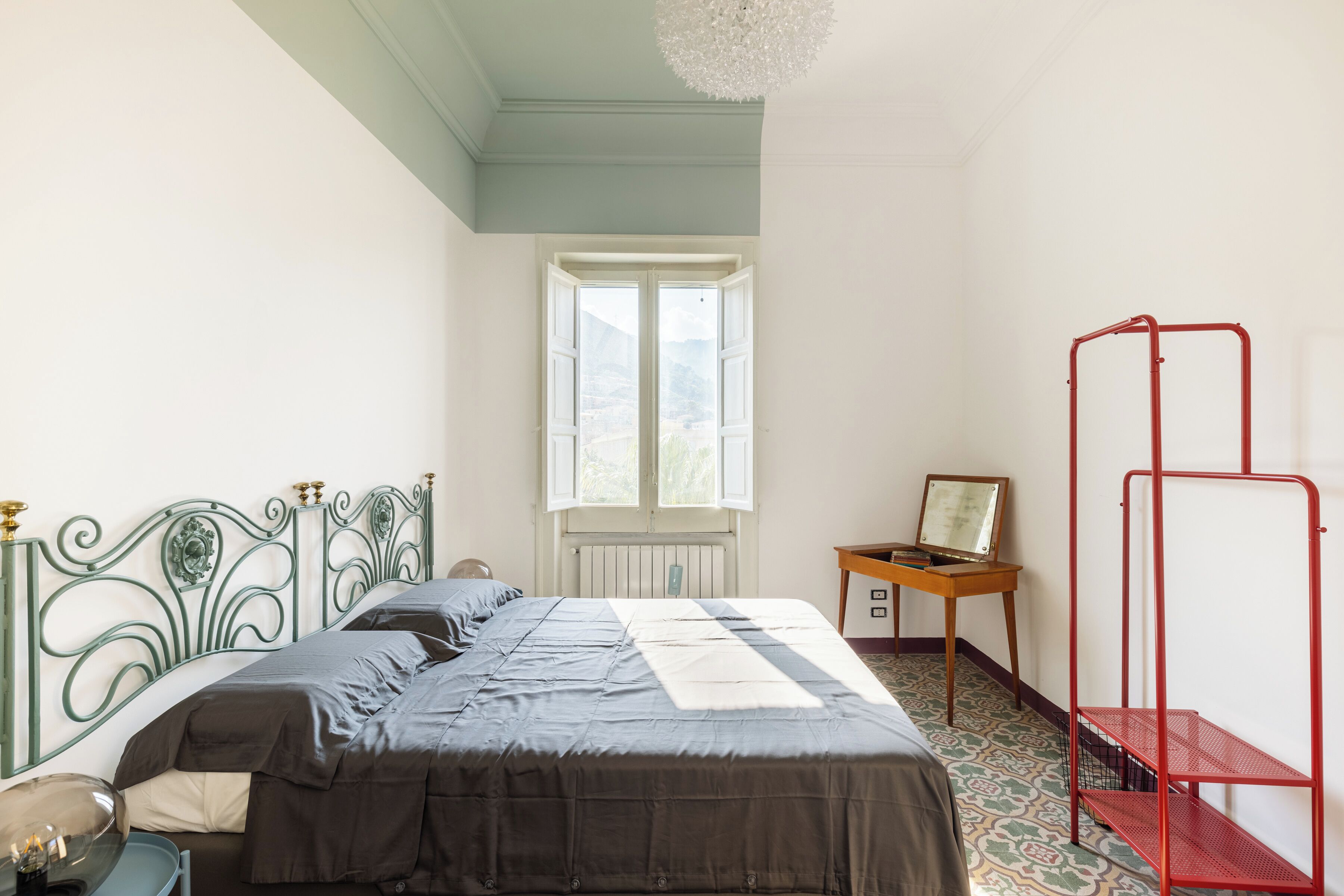 A bedroom with a bed, a window, a radiator, a desk, and a red metal shelf.