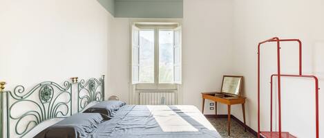 A bedroom with a bed, a window, a radiator, a desk, and a red metal shelf.
