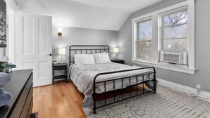 A bedroom with a metal bed frame, two bedside tables with lamps, a door, and a window with blinds. - Experience KC from Twin Bells: Two Charming Bungalows, One Prime Location! (Kansas City)