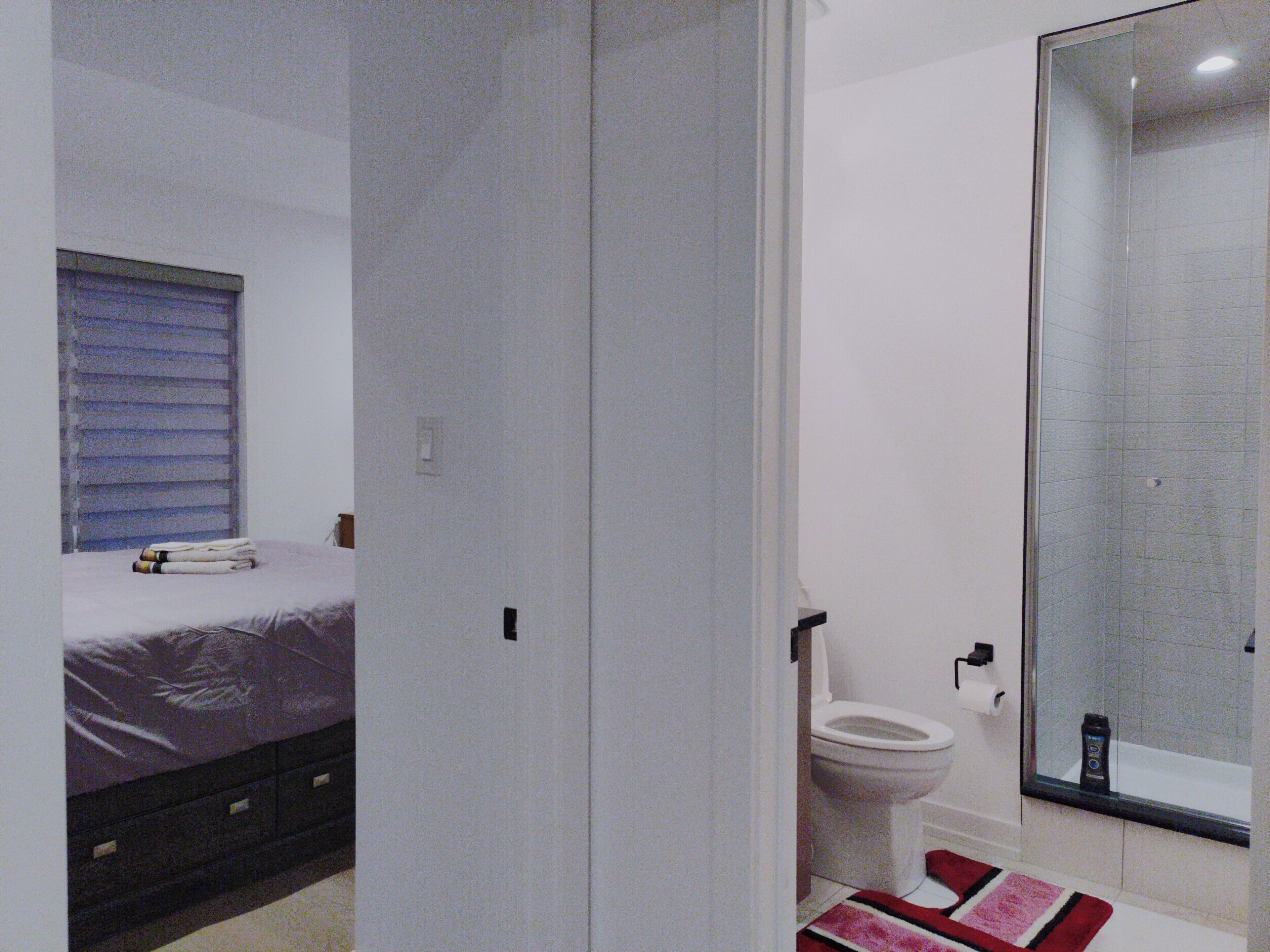 Ein Schlafzimmer mit Bett, ein Badezimmer mit Toilette und Dusche sowie ein Fenster mit Jalousien.