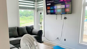 Ein modernes Wohnzimmer mit grauem Sofa, einem Standventilator, einem Wand-Fernseher, der verschiedene Apps anzeigt, und einem großen Fenster mit Jalousien.