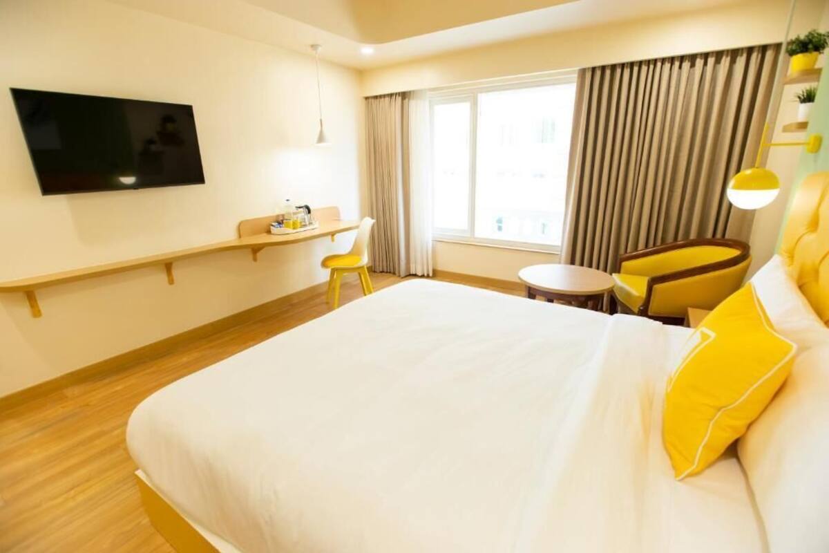 Una camera d'albergo con un letto, una TV fissata al muro, una scrivania piccola con una sedia e una finestra con tende.