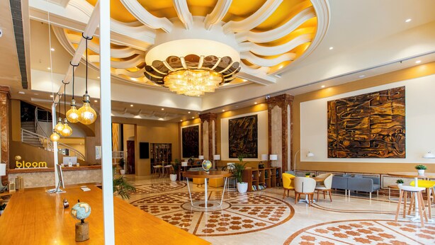 A lobby with a wooden reception desk, a globe, and a chandelier. There are seating areas with chairs and tables, and a large abstract painting on the wall. - Bloom Hub - Guindy (Chennai)