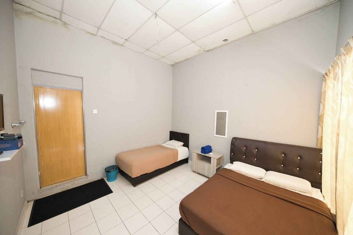 Ein Hotelzimmer mit zwei Betten, einer Holztür, einem kleinen Nachttisch mit Lampe und einem gefliesten Boden.