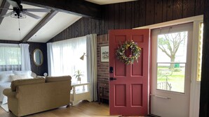 A cozy living room with a beige sofa, a red door with a wreath, and a view of a park through a window. - Pet-friendly, Kid-friendly Farmhouse near Holy Hill, Ice Age Trail, State Forest (Hubertus)