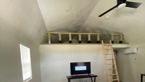 Una sala de estar con un sofá gris, un televisor sobre un mueble bajo, un ventilador de techo y una escalera de madera.