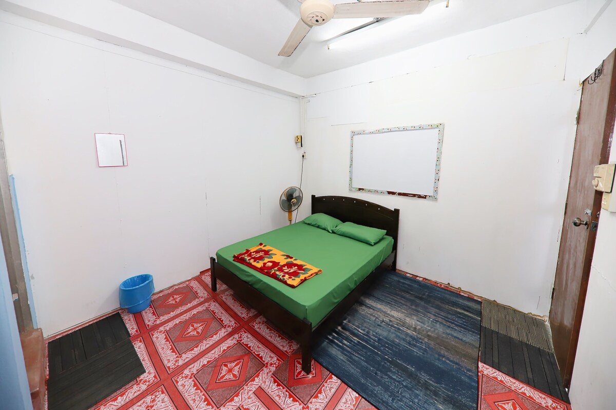 Een kleine, schone kamer met een enkel bed, een plafondventilator, een ventilator aan de muur en een deur met slot.