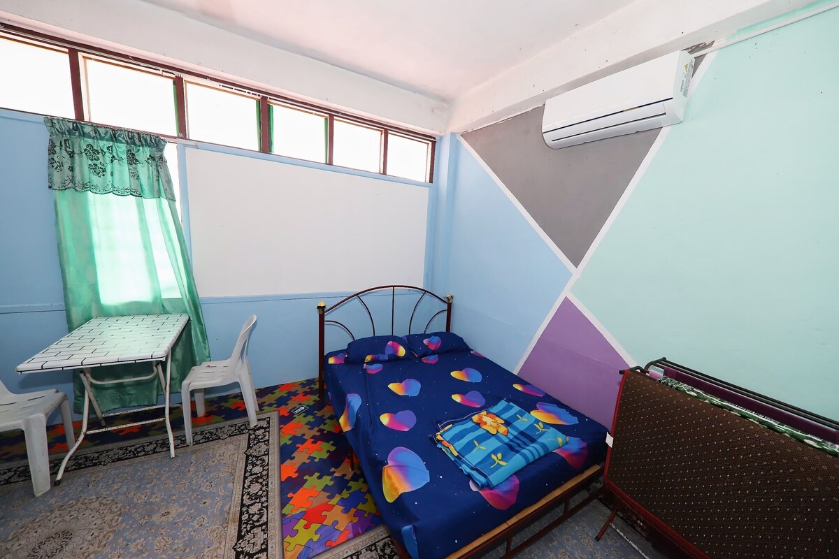 Een kleine eenpersoonskamer met een metalen bedframe, een tafel met stoel, een kamerbreed tapijt met patroon en een airconditioningunit aan de muur.