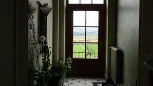 Un couloir doté d’un sol carrelé, d’une grande fenêtre donnant sur des champs, d’un luminaire suspendu et de plantes en pot.