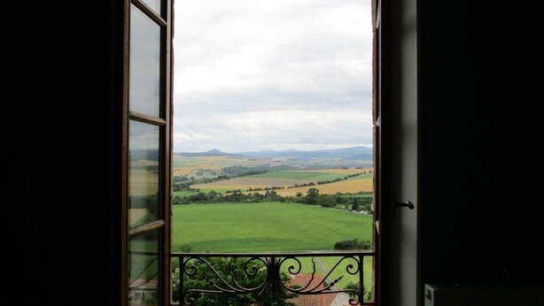 Une vue depuis une fenêtre, avec un balcon en métal, donnant sur un paysage verdoyant composé de champs et de collines lointaines.