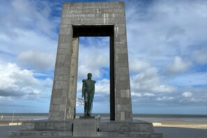 A monument with a statue in the center, surrounded by a stone archway, set against a beach and ocean backdrop.