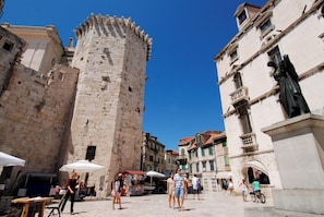Una torre in pietra storica e edifici in una piazza, con posti a sedere all'aperto e ombrelloni.