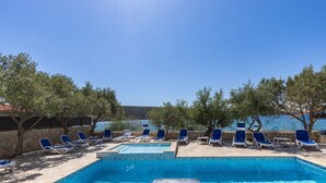 A swimming pool with blue tiles, surrounded by lounge chairs and trees, with a clear view of the sea in the background. - Beachfront Santa Maria 2 Apartment 1 (Novalja)