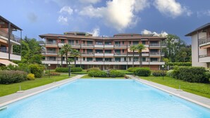 Una piscina rodeada de un césped cuidado, con un edificio de varios pisos y balcones al fondo.