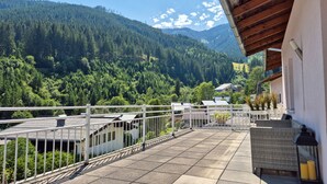 A balcony with a view of a forested mountain, a white railing, and a building with a tiled roof. - Penthouse Summer Winter Fun 4 Roof Terrace (Zell am See)