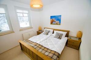 Een slaapkamer met een houten bed, wit beddengoed, een geruit plaid, een nachtkastje met een lamp, en een schilderij van honden aan de muur.
