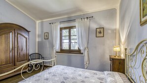 A bedroom with a bed, a wooden wardrobe, a chair, a window with curtains, and a bedside table with a lamp. - Maramaldo Galestro Shared Pool Chianti (Castelfiorentino)
