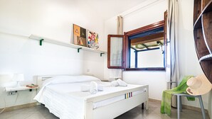 Une chambre à coucher comprenant un lit, une table de chevet, une chaise, une fenêtre et une étagère avec des œuvres d’art.