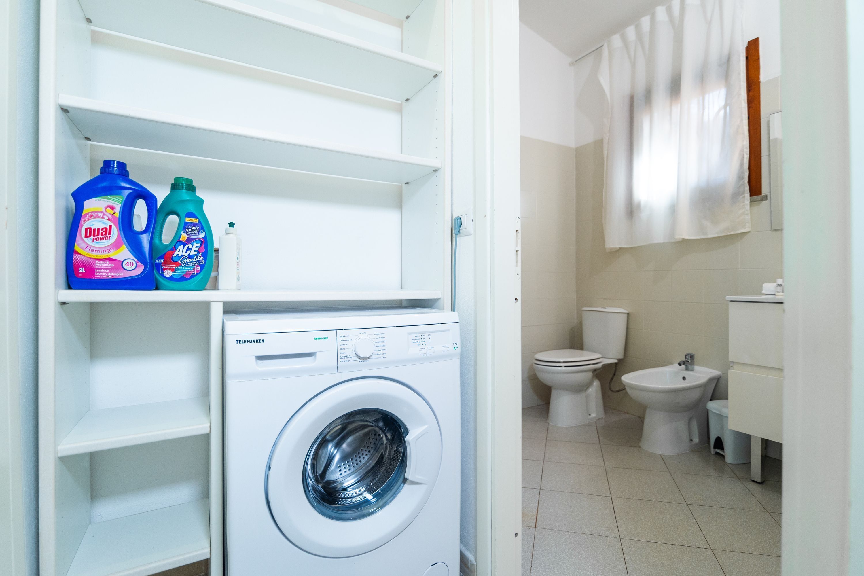 A laundry area with a washing machine, shelves with detergent bottles, a toilet, bidet, and a sink.
