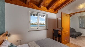 Une chambre avec un plafond en bois, un lit recouvert d’une couverture blanche et grise, un bureau avec une chaise et une fenêtre donnant sur une plage et des montagnes.