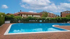 Ein Schwimmbecken mit Ziegelumrandung, umgeben von einem Zaun, mit Häusern im Hintergrund.