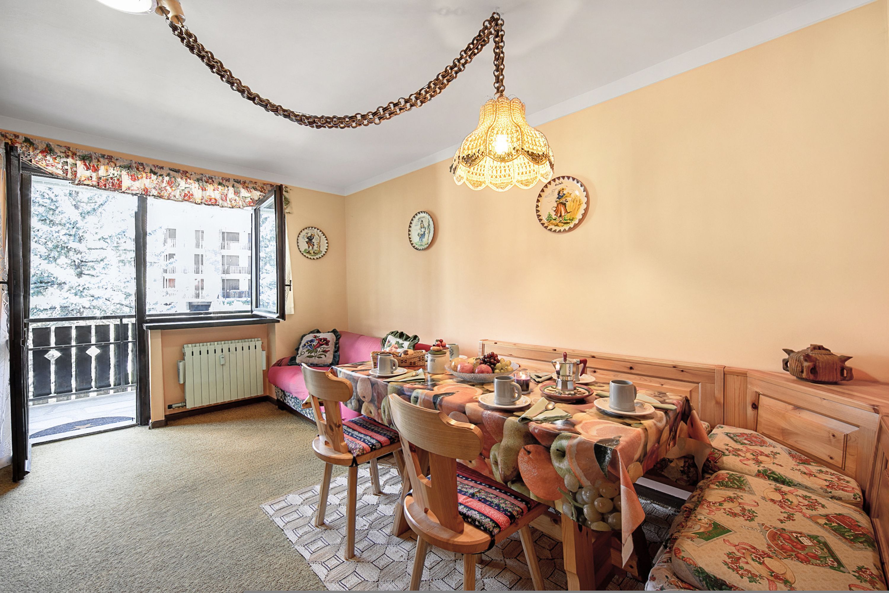 A cozy dining area with a wooden table set for four, surrounded by chairs with patterned cushions. A decorative hanging light fixture is above the table, and a bench with a floral cushion is against the wall.