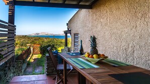 Une table en bois, sur laquelle se trouvent un panier de fruits, une bouteille de vin et des verres, est placée sur une terrasse offrant une vue sur la mer et les montagnes.