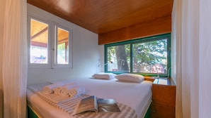Een slaapkamer met een bed, kussens, een nachtkastje, een raam met uitzicht op bomen en een houten plafond.