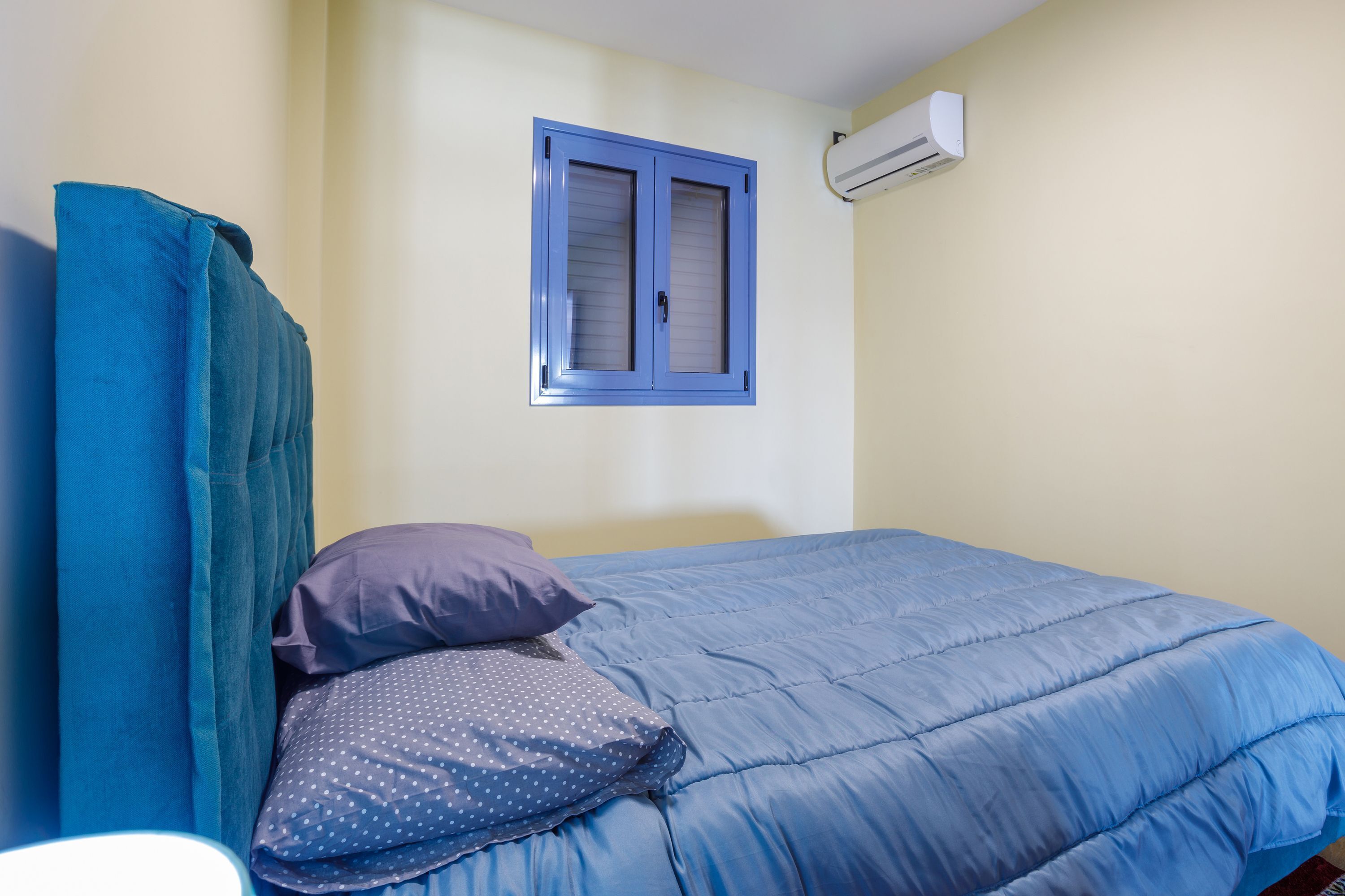 Une chambre à coucher avec un lit, une fenêtre avec des volets bleus et un appareil de climatisation fixé au mur.