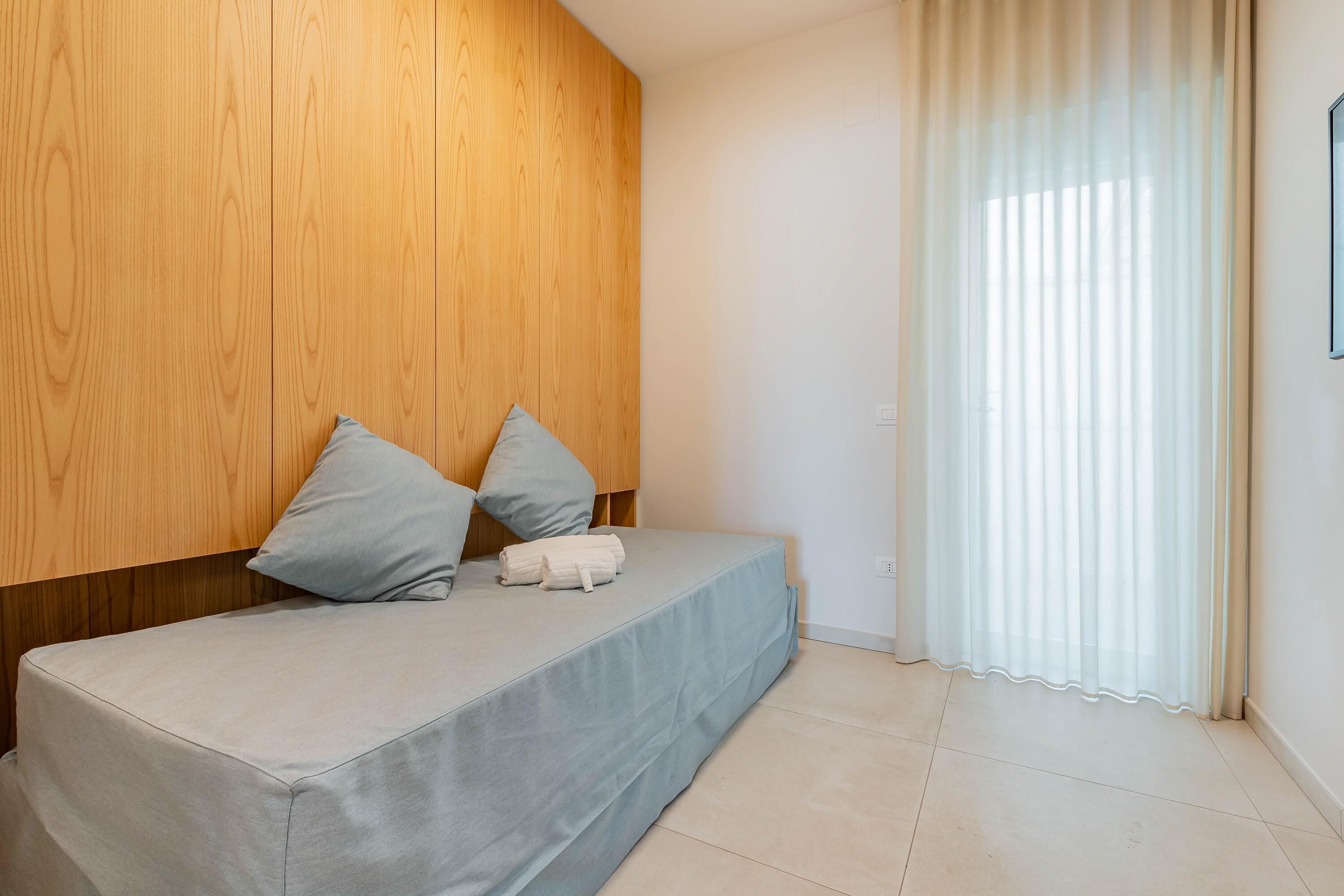 A modern bedroom with a wooden headboard, a bed with a grey mattress and pillows, and a window with sheer curtains.