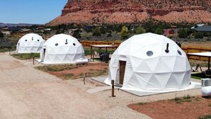 Three dome-shaped structures with a geometric pattern, set against a backdrop of a red rock formation and a clear blue sky. - White Camel (Kanab)