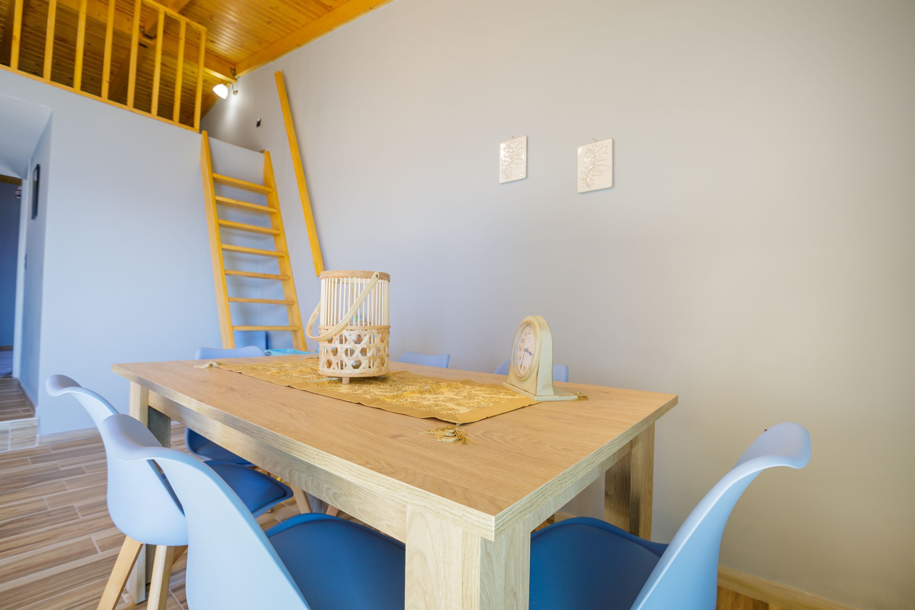 Un coin repas avec une table en bois, des chaises bleues, une échelle en bois et une horloge fixée au mur.