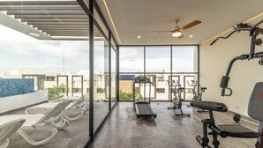 A spacious gym with exercise equipment, large windows, and a view of the cityscape. - Stylish Boho Studio + Rooftop Pool @ Aldea Zama (Tulum)