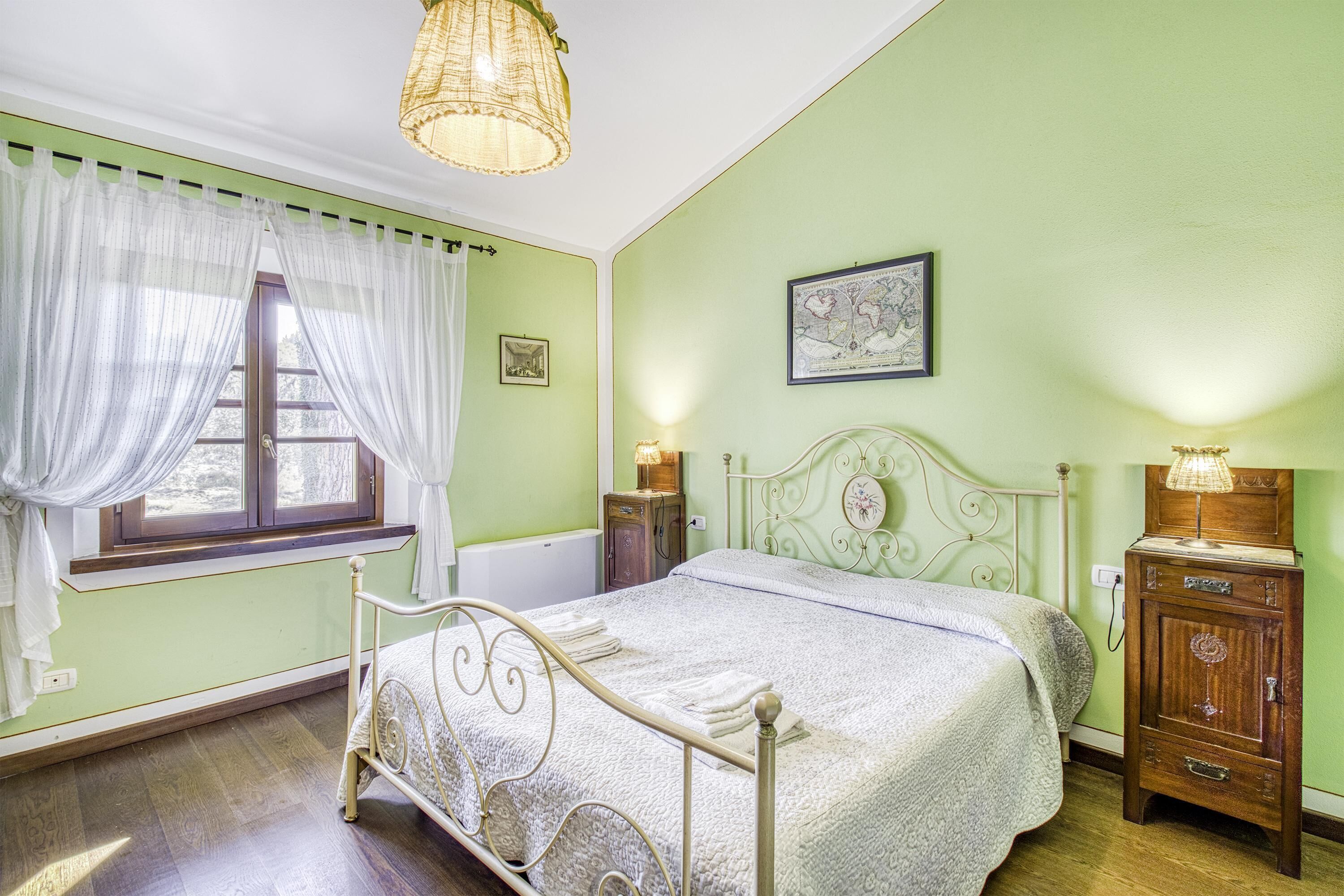 Un dormitori amb un llit, una tauleta de nit, una petita amagatzematge, una finestra amb cortines, una lluminària penjant i un mapa fixat a la paret.