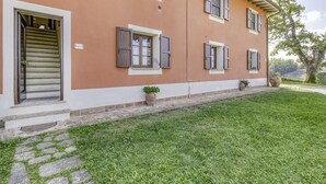 A two-story building with a brown facade, white trim, and shutters on the windows. There is a stone pathway leading to the entrance, and potted plants are placed along the pathway. - Maramaldo Sangiovese Shared Pool Chianti (Castelfiorentino)